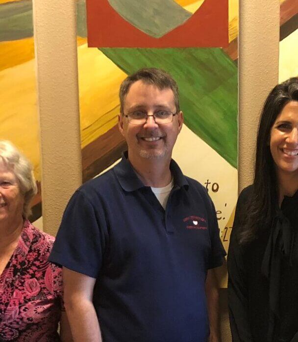 Light skin toned man with ash grey hair standing next to two other women wearing a navy blue collared shirt.